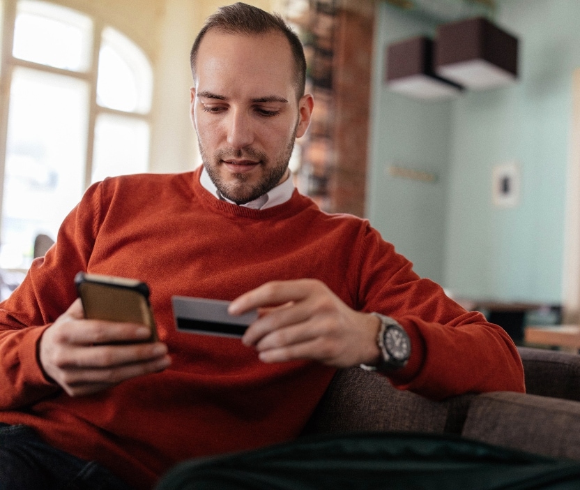 A man sits on a couch with his phone in one hand and his credit card in the other.