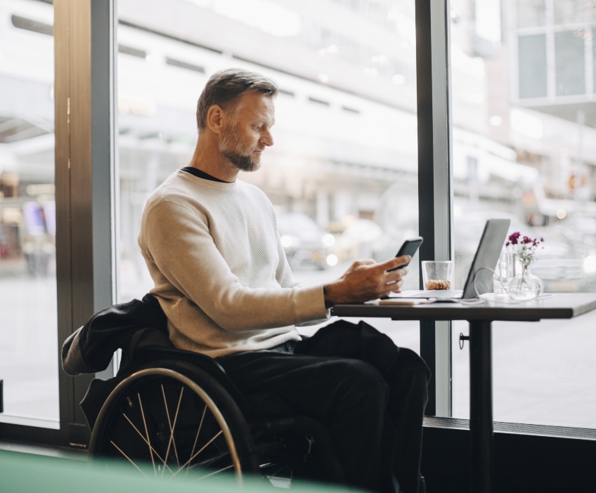 A man in a wheelchair sits in a café window while looking at his cellphone and worrying about romance scams.