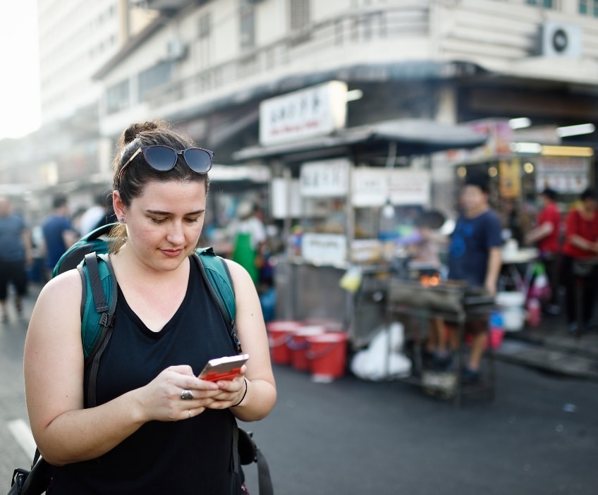 A woman stands in the street in Asia with a backpack on, looking down at her phone.