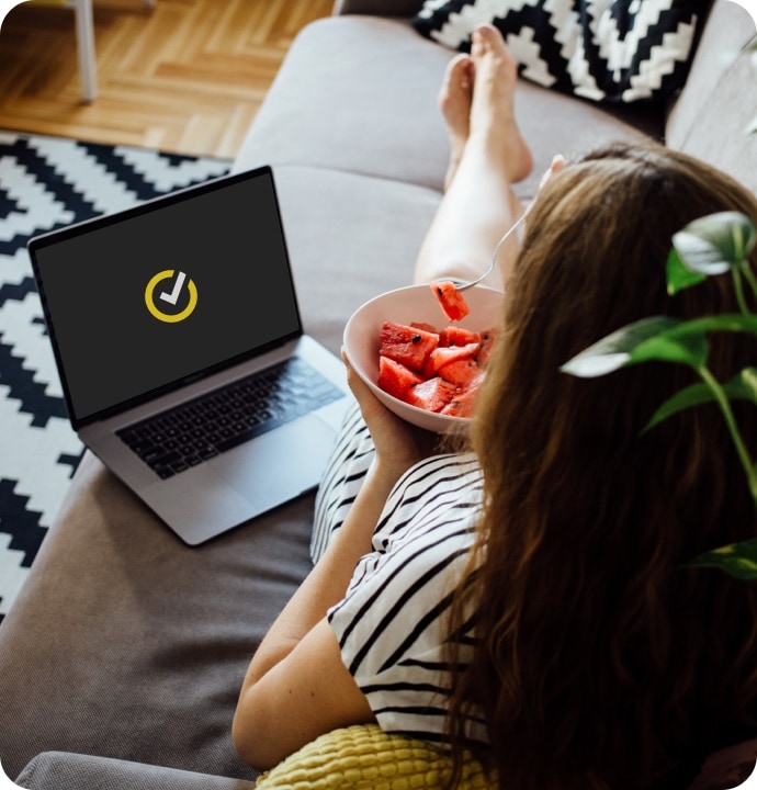 Woman eating fruit with laptop beside her displaying NortonLifeLock symbol.