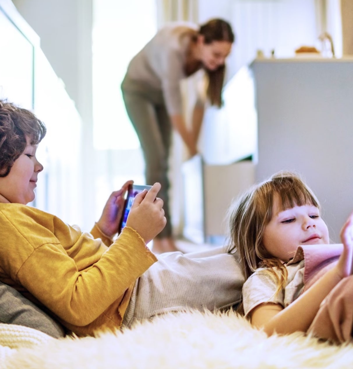 Kids lying down using tablet devices while mother in the background is tidying the kitchen.